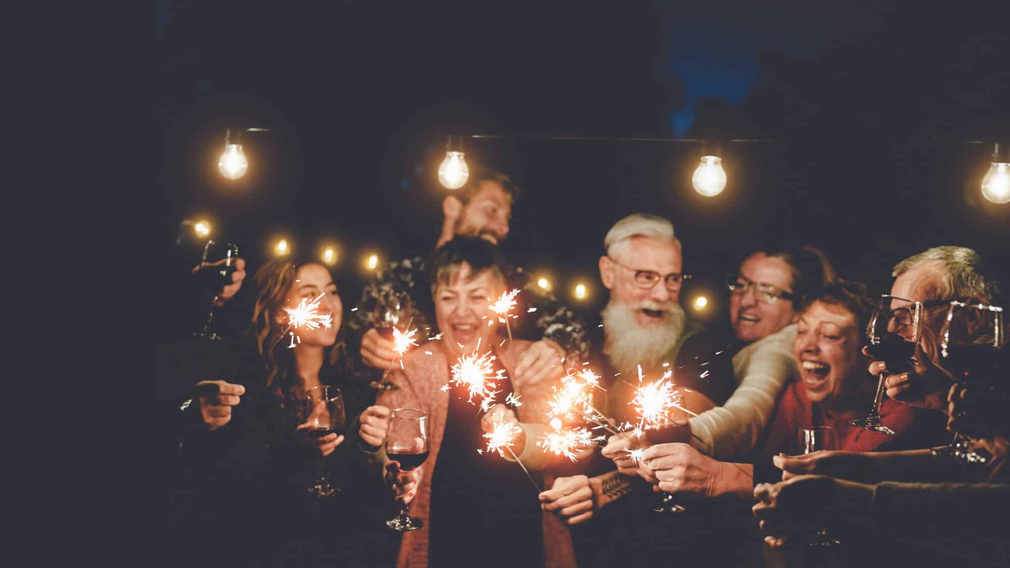 Happy family having at dinner party outdoor - Group of multiracial older and young people celebrating together drinking wine holding fireworks sparklers - Concept of youth and elderly parenthood