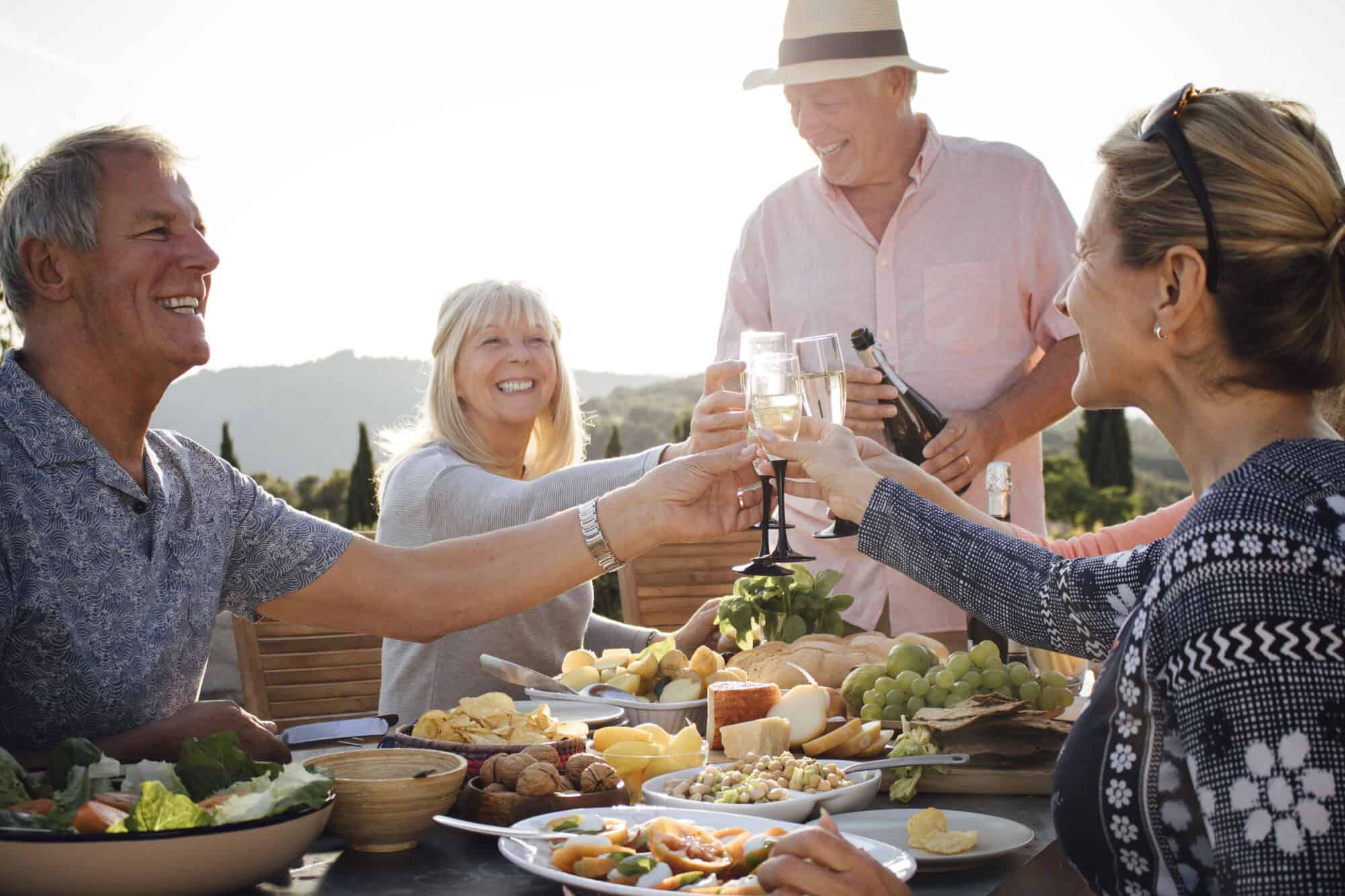 A group of mature friends are sitting around an outdoor dining table, eating and drinking. They are cheering with a glass of champagne and enjoying each others company. The image has been taken in Tuscany, Italy.